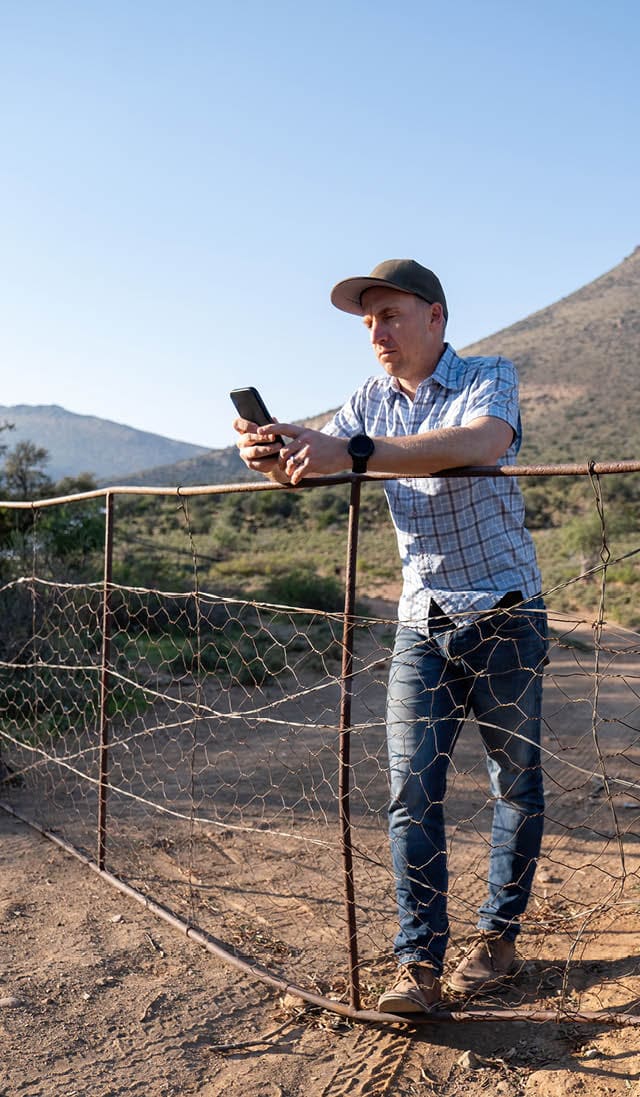 A farmer takes a break while in the field, he checks his mobile phone while he leans against an old fence.