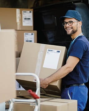 Young happy manual worker carrying cardboard boxes in delivery van while communicating with his colleagues.